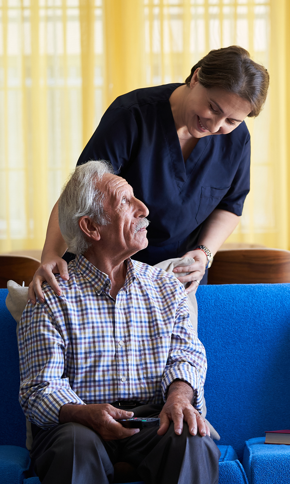 caregiver-and-patient-in-home patient sitting on blue couch with caregiver standing checking on them